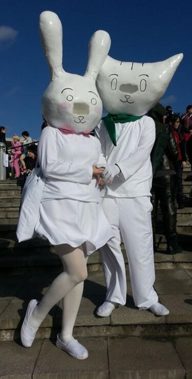 A pair of cosplayers stood on concrete steps wearing white clothing and headpieces in the style of the cat and rabbit from web animation There She Is!!! by SamBakZa