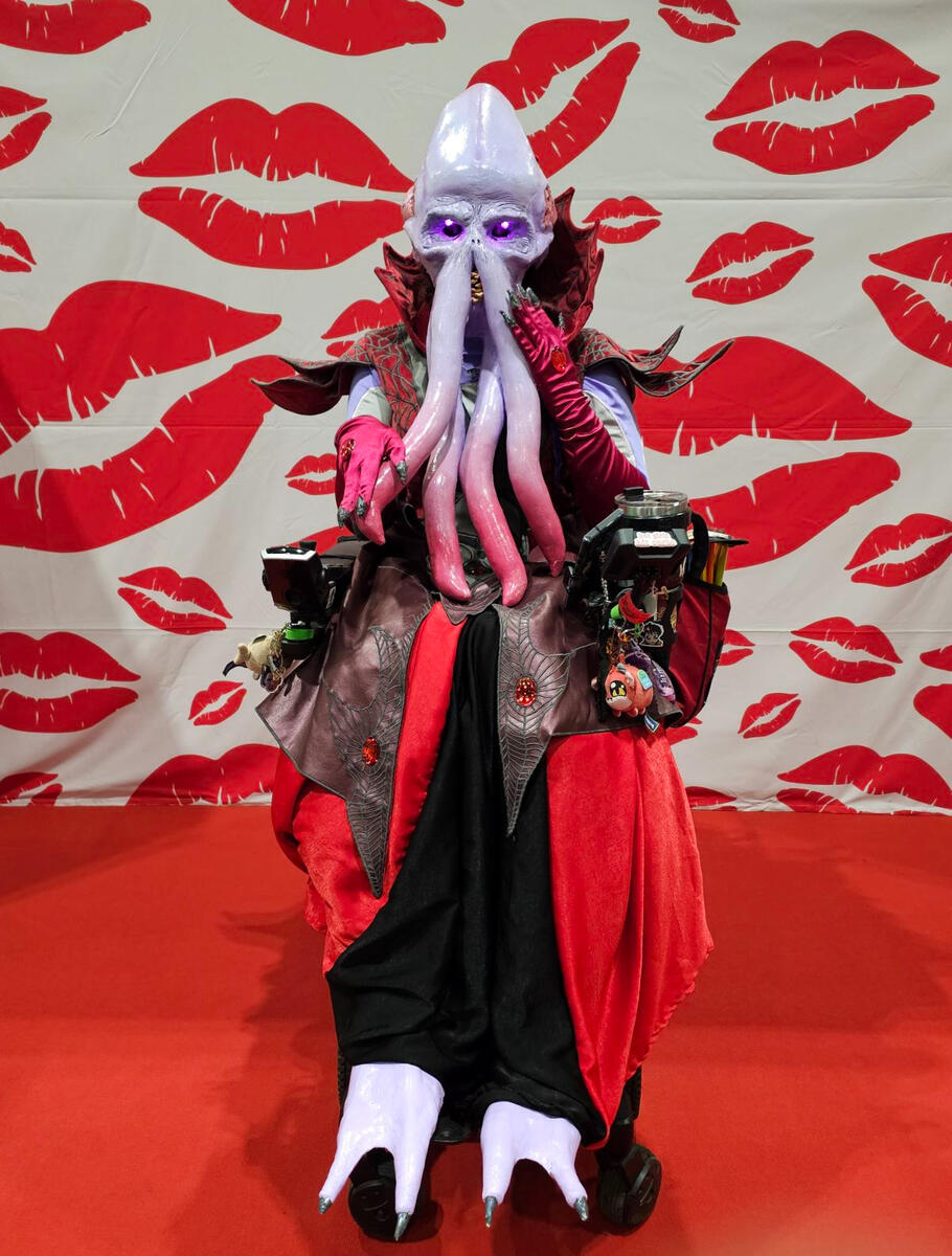 A cosplayer in a powered wheelchair sat in front of a backdrop of lipstick kiss patterns