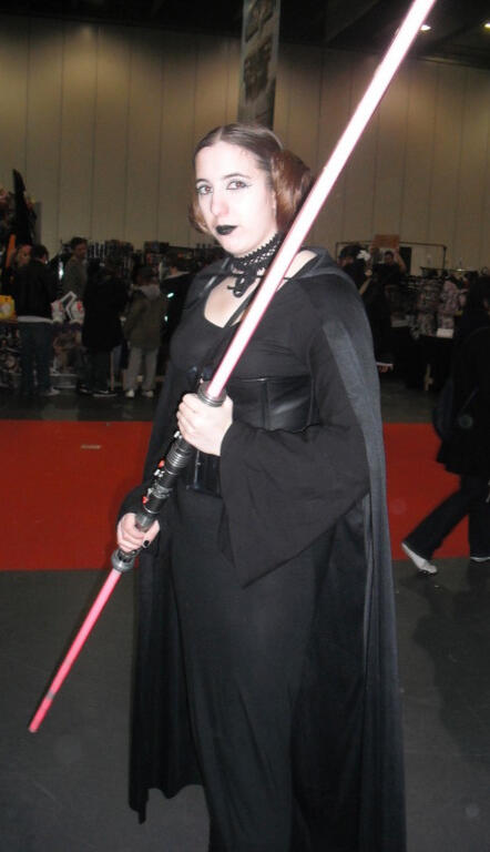 A cosplayer dressed like Princess Leia except the costume is all black with a leather underbust corset, black cloak, black lipstick, and a double bladed red lightsaber.