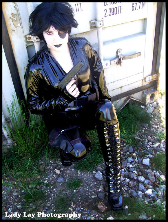 A cosplayer in a black PVC catsuit and short black wig with pale facepaint and a black mark over one eye, holding a plastic prop gun and crouching by a shipping container.