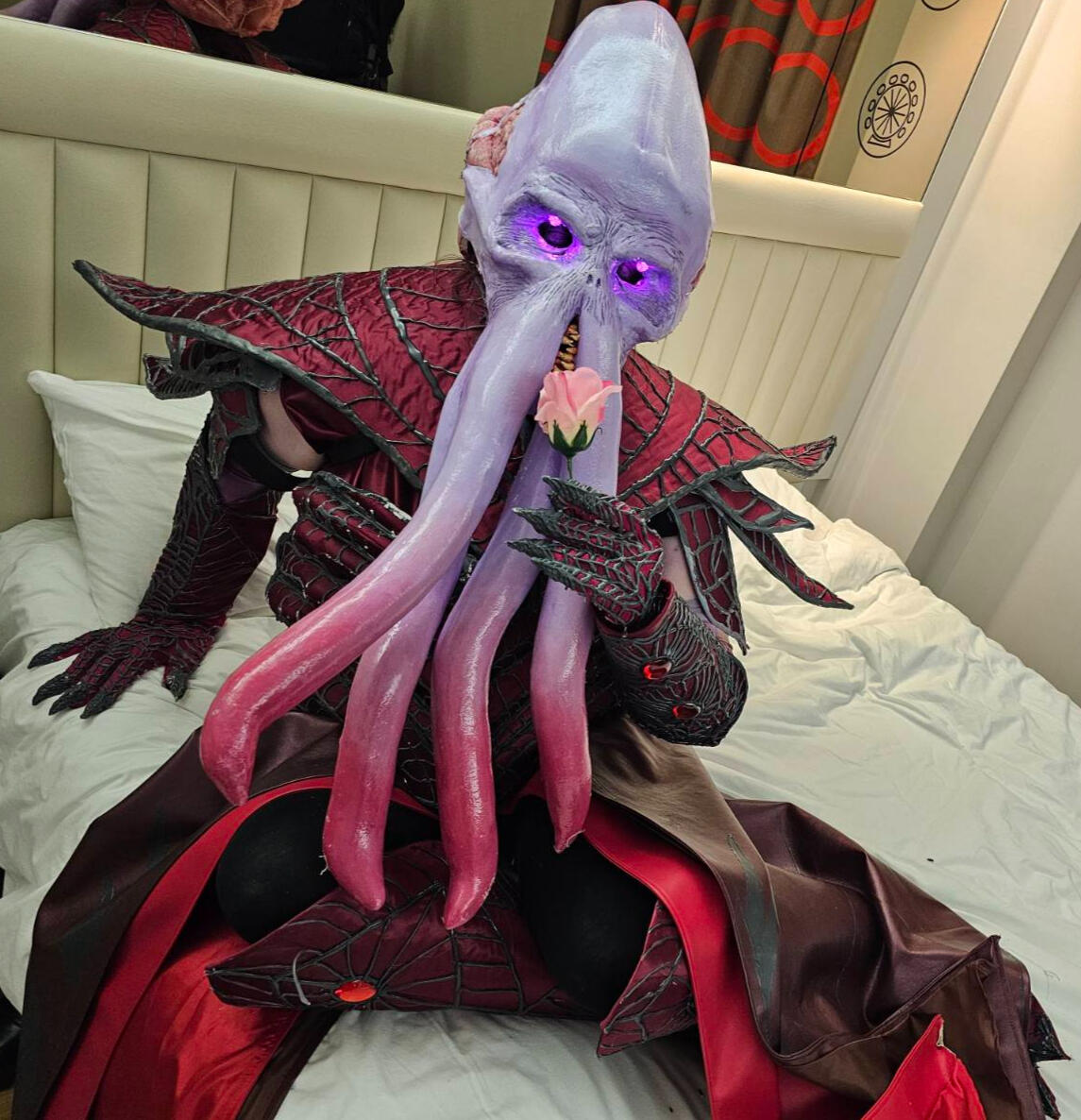 A cosplayer wearing an illithid head mask with glowing eyes and red and grey armour matching the style of The Emperor from Baldur's Gate 3.