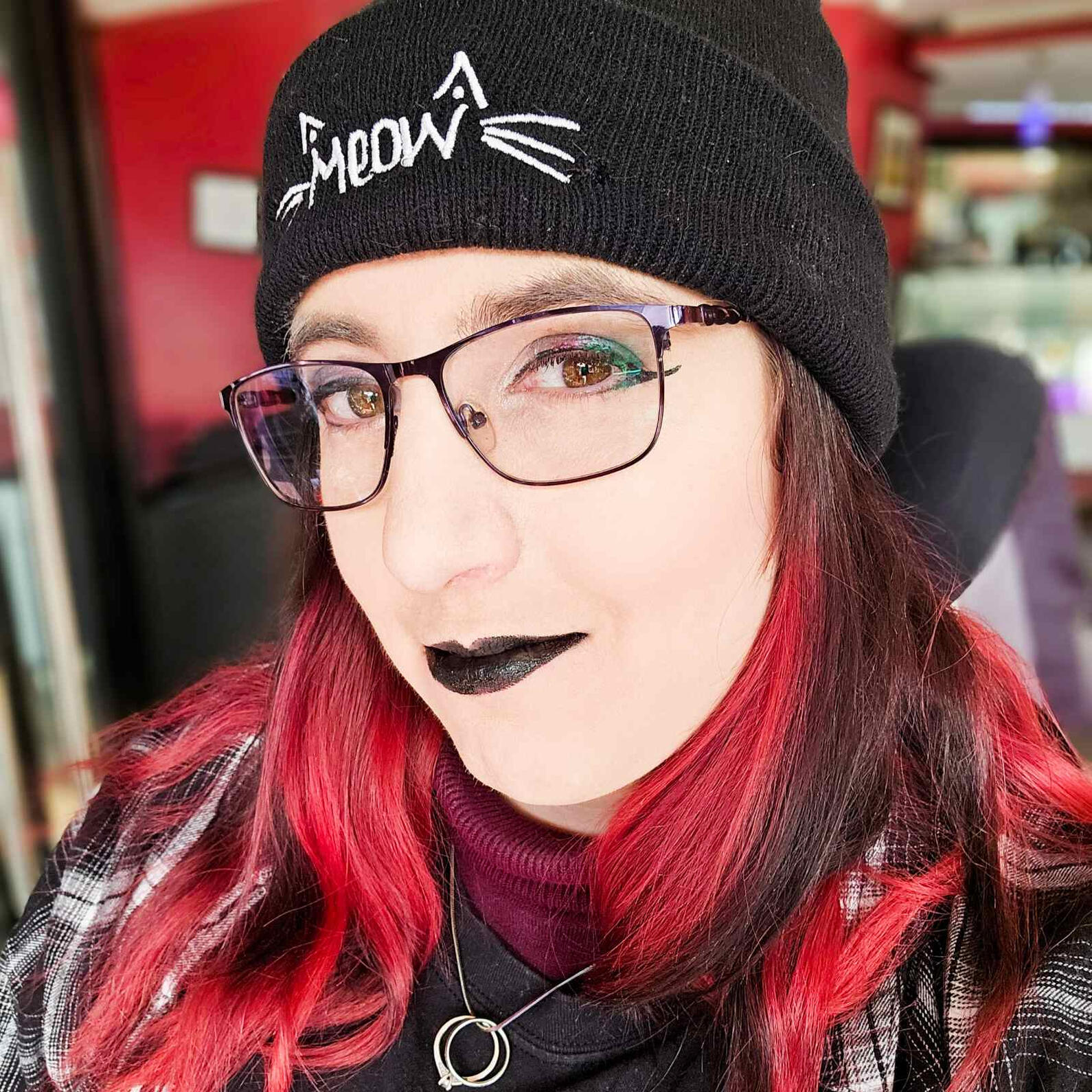 A woman with red and black hair wearing a black knitted hat with the word Meow embroidered on the front. She is also wearing glasses and goth style makeup.