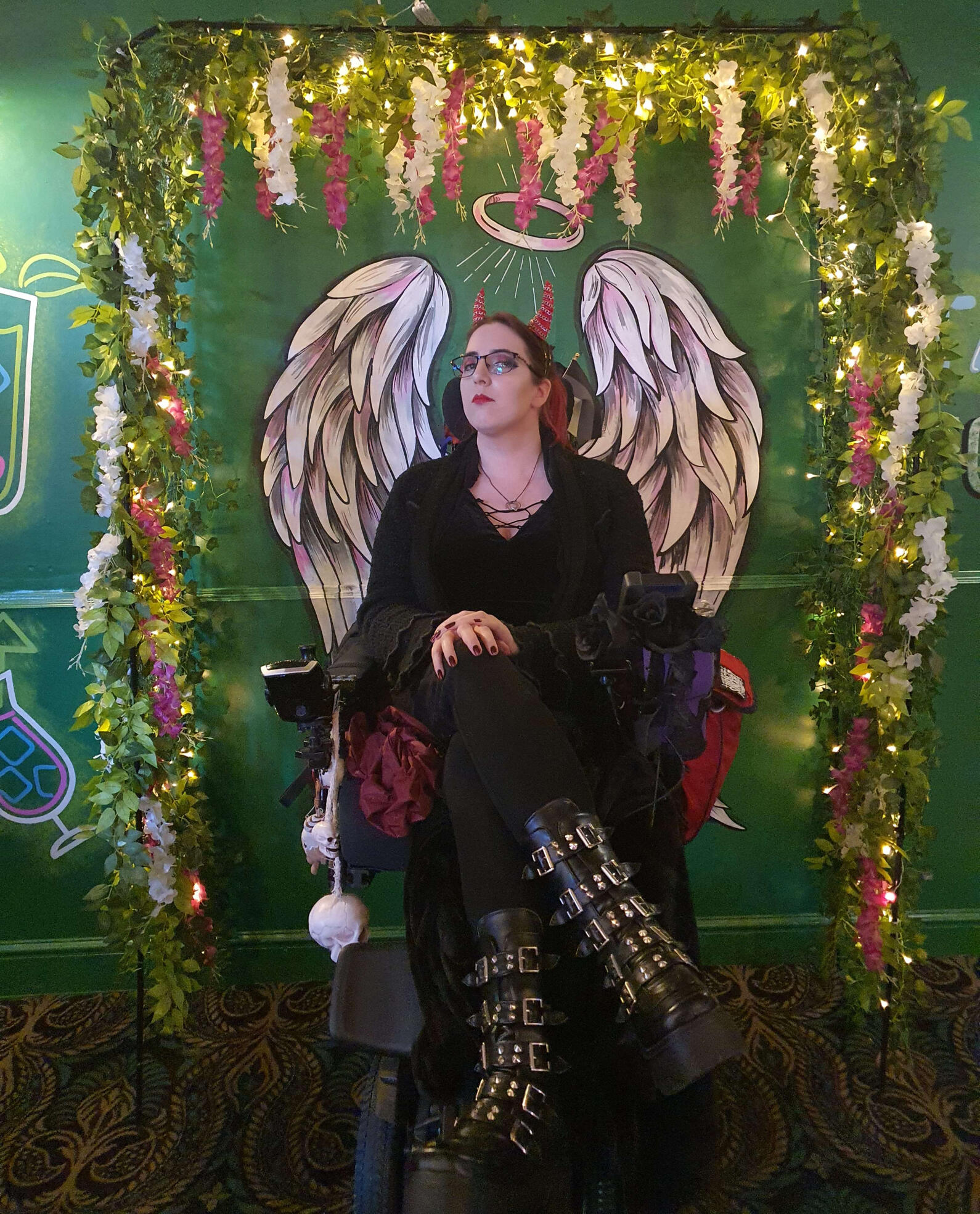 My...Me! A woman with red and black hair wearing an all black outfit with tall platform boots with buckles and spikes. She has a red devil horn headband and is sat in a powered wheelchair in front of a wall that has angel wings and halo painted on it and a lit arch