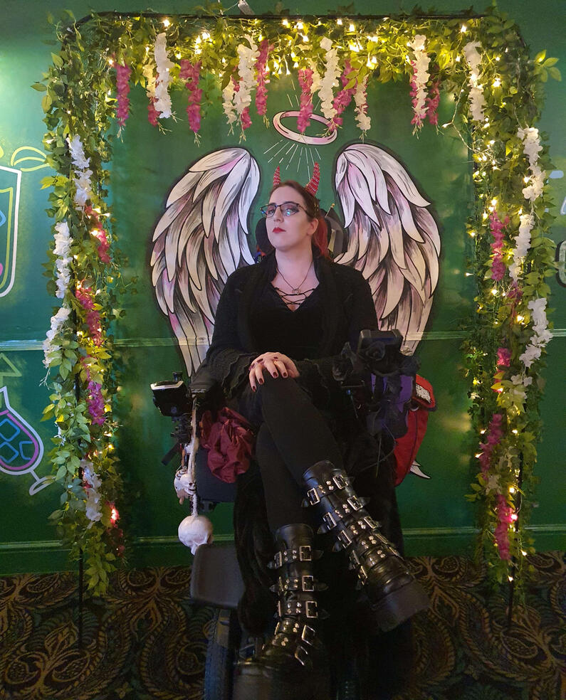 My...Me! A woman with red and black hair wearing an all black outfit with tall platform boots with buckles and spikes. She has a red devil horn headband and is sat in a powered wheelchair in front of a wall that has angel wings and halo painted on it and a lit arch